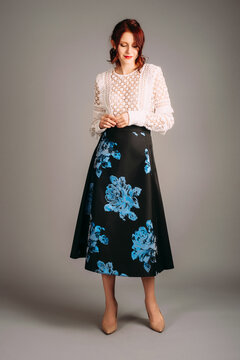 Long sleeve blouse in white and midi skater skirt in black. Studio portrait of shy young woman on high heels. Female evening outfit. Ginger lady smiling on studio background.