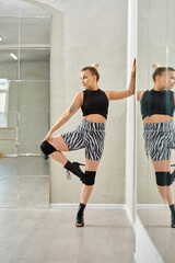 fashion-forward woman in zebra shorts and black top balances on one leg while dancing against mirror