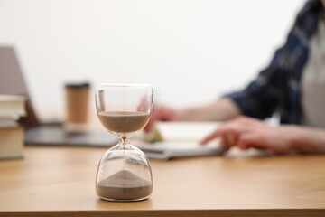Hourglass with flowing sand on desk. Man taking notes indoors, selective focus
