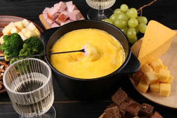 Fondue pot with melted cheese, fork and different products on black wooden table