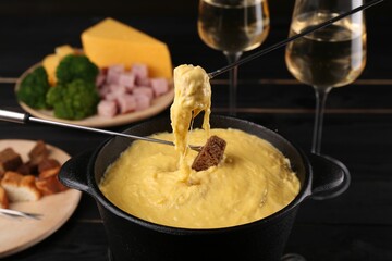 Dipping pieces of ham and bread into fondue pot with melted cheese on table, closeup