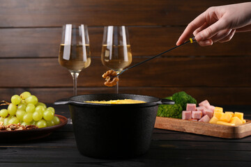 Woman dipping walnut into fondue pot with melted cheese at black wooden table, closeup