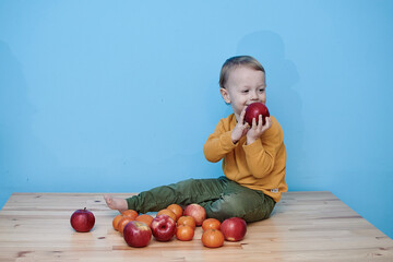 Fototapeta premium a little boy of 2 years old sits on a table against a blue wall and holds a red apple. there are a lot of red apples and a lot of tangerines around. child in green pants and yellow sweater