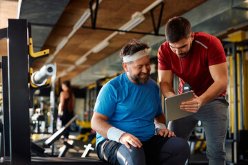 Happy mature athlete and his coach using touchpad during exercise class in gym. - obrazy, fototapety, plakaty