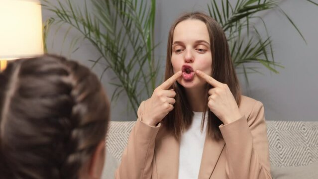 Language correction and therapy. Sound development in speech. Confident teacher speech therapist practicing pronunciation exercises with little girl child while sitting on sofa