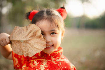 A joyful young girl in a red cheongsam playfully hides behind a large dried leaf, outdoors on a...
