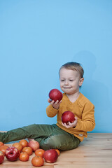 Fototapeta premium a little boy of 2 years old sits on a table against a blue wall and holds a red apple. there are a lot of red apples and a lot of tangerines around. child in green pants and yellow sweater