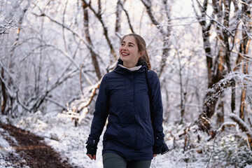 Cheerful hiker girl in outdoor gear walking through a magical snowy forest in the mountains on a sunny day, young woman looking up and admiring nature while out hiking in the winter woods, tourism 