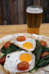 Closeup view of a pizza with mozzarella cheese, spinach, cherry tomatoes and fried eggs, on the wooden table.
