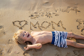 cheerful boy 5-6 years old son having fun on beach. heart is drawn on the sand and the word mom is written. Positive human emotions, feelings, joy, happy childhood. gift from a child for Mother's Day