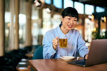 An Asian woman drinks tea at the cafe while working on her laptop.