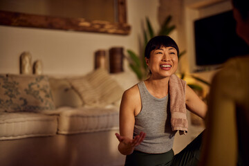 Focus on the smiling Asian yoga instructor, sitting on the floor, talking to a female client.