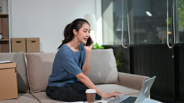 Asian woman with a smile standing holding notepad and tablet in living room on the sofa at home office.