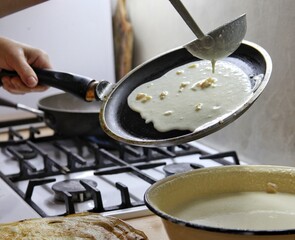 cooking pancakes in a frying pan at home, first person and side view. a frying pan in the hands of a woman and in the other hand a ladle with dough for pancakes