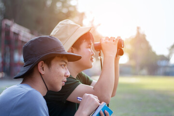 Asian teenboys learning nature by using binoculars to watch birds and insects in public park during summer camp of their school, idea for learning creatures and wildlife animals outside the classroom.