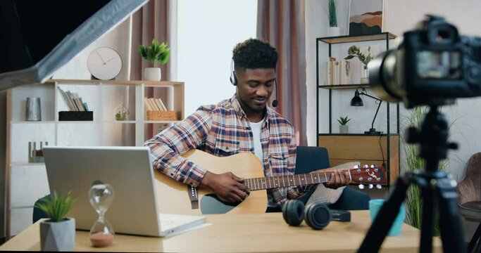 Handsome joyful man in headphones sitting in front of camera at home and recording videoblog for his social media followers on guitar play theme