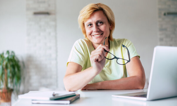 Portrait of cute smiling mature woman while she is looking on camera and holding glasses in hands during break after work on laptop on home office