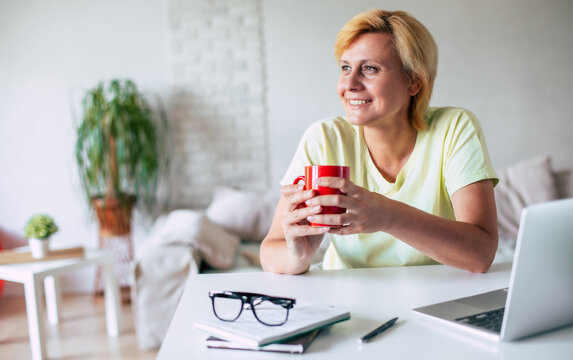 Image of happy calmed mature woman in casual clothes with beverage in hands looking away while sitting at a desk in the kitchen at home.