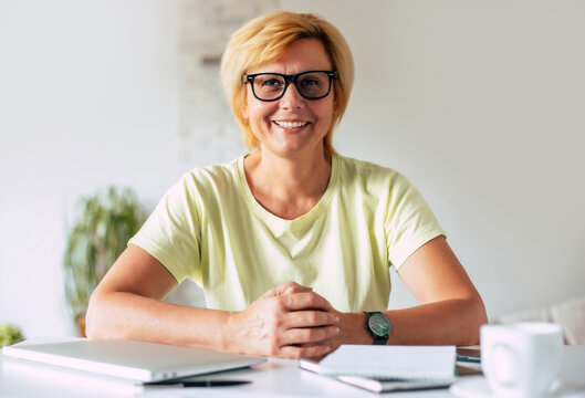 Smart confident smiling mature woman wearing eyeglasses is looking on camera while sitting near work desk at home office. Freelancer and retired person