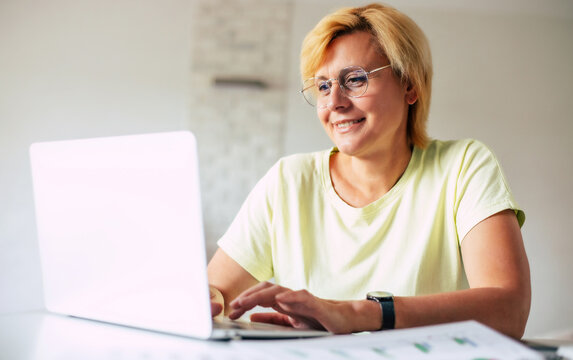 Side view shot of smart confident happy mature woman wearing goggles using laptop while working remotely from home in living room or making some purchase online from home