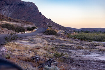 Empty rural black asphalt road between arid terrain with rocky mountains and yellow curved road sign in background, sunset in Baja California Sur desert, Mexico