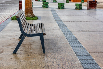 Empty wooden bench on sidewalk of promenade, street and small green pillars in background, cloudy...