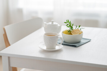 A cup of tea with lemon and a teapot on a white table against the background of a kitchen window.