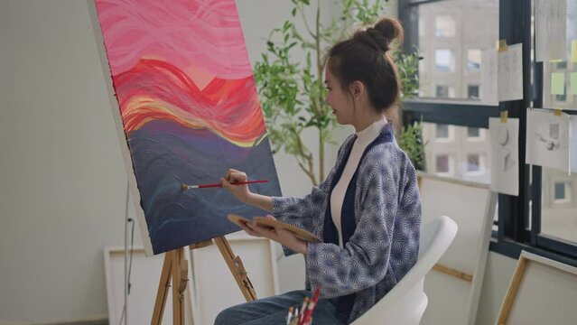 happy asian female painter using red paint on canvas,holding tube of oil paint,fine art artist drawing,asian woman painting brush on canvas.concept of enjoy leisure time with art hobby in art studio