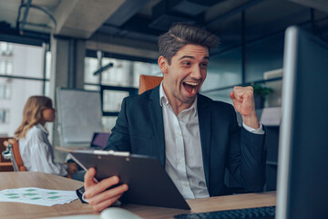 Excited joyful male manager holding clipboard and making winner yes hand while sitting in office