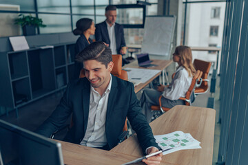 Happy male manager sitting at desk with clipboard and thinking about project while looks on computer