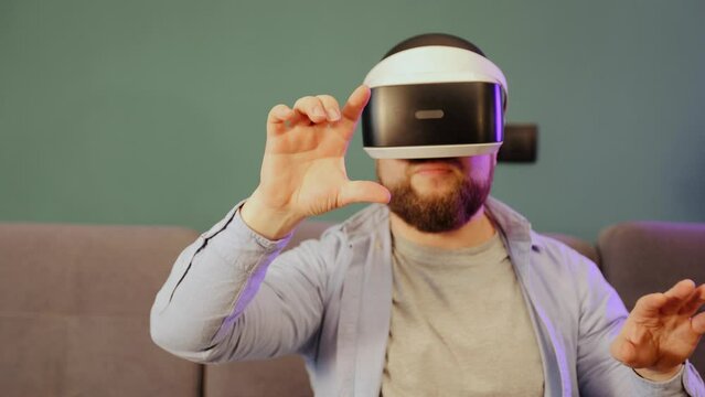 Close-up portrait of a Caucasian young man wearing VR glasses, moving his hands in the air and making some gestures like scaling and swiping photos in virtual reality in his living room at home