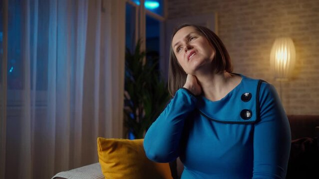 Neck pain, woman suffering from ache at home, health problems concept. Middle aged woman suffering from neck pain sitting on sofa at home. unhappy female suffering from neck pain at home