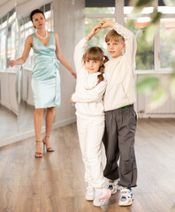 Motivated preteen ballroom dancers, girl and boy in sportswear practicing elegant dance moves in pair in bright studio, with female coach supervising in background..