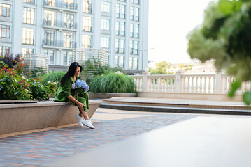 Adult brunette woman in green fashion costume sitting on bench at urban city square, with hydrangea...