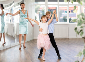 Boy and girl kids repeat movement while taking rumba dancing lessons. Female teacher conducts class for students during dance workout and watches performing movements