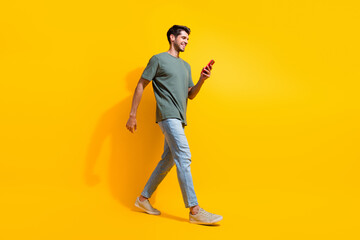 Fototapeta na wymiar Full length photo of cool positive guy wear khaki t-shirt walking communicating modern device isolated yellow color background