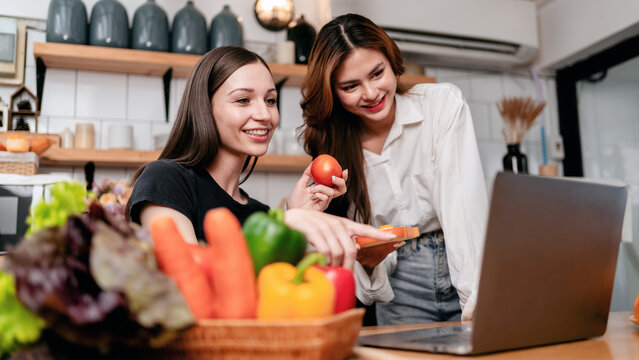Two young woman learning and watching food recipe tutorial on laptop together while cooking healthy meal