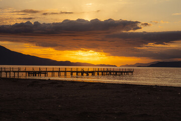 Fototapeta premium A dramatic sunset in the Mediterranean. The sun behind the clouds, beach and a dock in the foreground. 