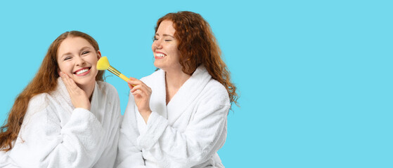 Happy redhead women in bathrobes and with makeup brush on blue background with space for text