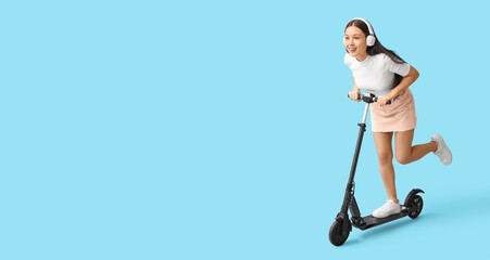 Beautiful young woman in headphones riding modern electric kick scooter on blue background with space for text © Pixel-Shot