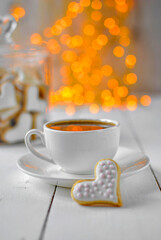 A cup of coffee. Next to the shortbread cookies in the shape of a heart. In the background there is a glass jar with cookies and yellow electric lights. Valentine's Day. Soft focus.