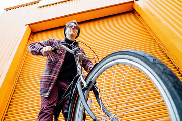 Bottom view Young stylish hipster man standing with a cruiser city Bicycle near orange Wall....
