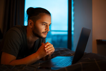 Serious young guy lying on bed and working home on laptop. Looking at the screen.