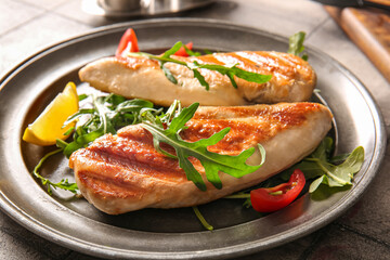 Plate of tasty grilled chicken breast with arugula, tomatoes and lemon on grey tile background