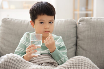 Ill little Asian boy with glass of water and pill sitting at home