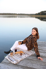 romantic vacation near the lake. Picnic on a wooden pier by the lake with food, croissants, coffee, chocolate and candles. aesthetic. the girl sits on a wooden pier near the lake and drinks coffee