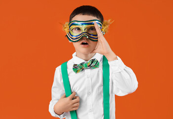 Surprised little boy in costume and carnival mask on orange background