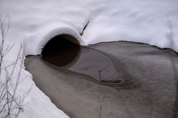 round pipe under the bridge and partially frozen ditch in winter. Snow background with water.