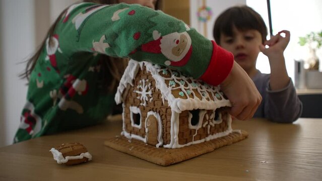 Children taking apart gingerbread home covered in royal ice, siblings brother and sister eating sugar bread