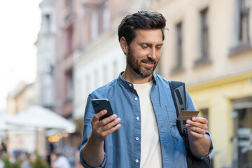 Mature bearded tourist with credit card and smartphone on evening city street - obrazy, fototapety, plakaty